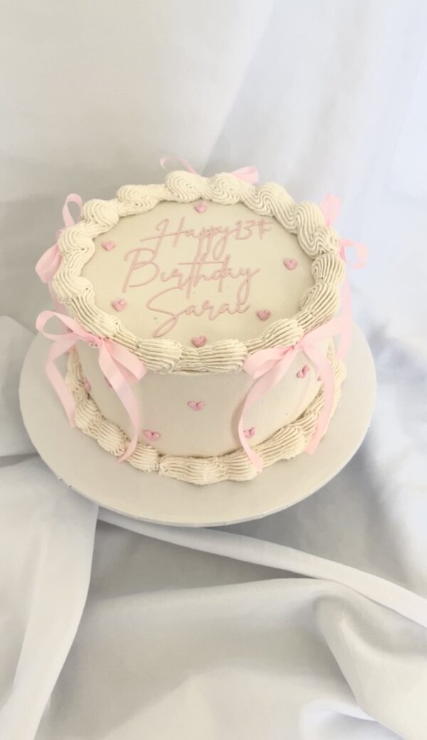 Angled view of vintage pink coquette-style buttercream cake adorned with pink bows, heart accents, and a cursive 'Happy Birthday' message, elegantly displayed on a white cake stand