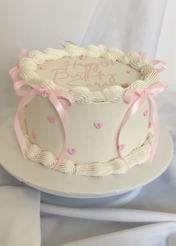 Side view of vintage pink coquette-style buttercream cake with pink bows and heart accents, featuring "Happy Birthday" message elegantly displayed on a white cake stand with a soft white backdrop.