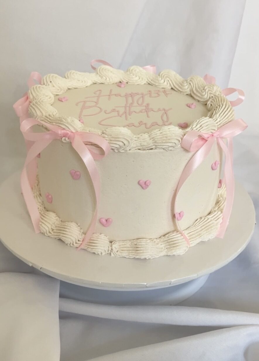 Side view of vintage pink coquette-style buttercream cake with pink bows and heart accents, featuring "Happy Birthday" message elegantly displayed on a white cake stand with a soft white backdrop.